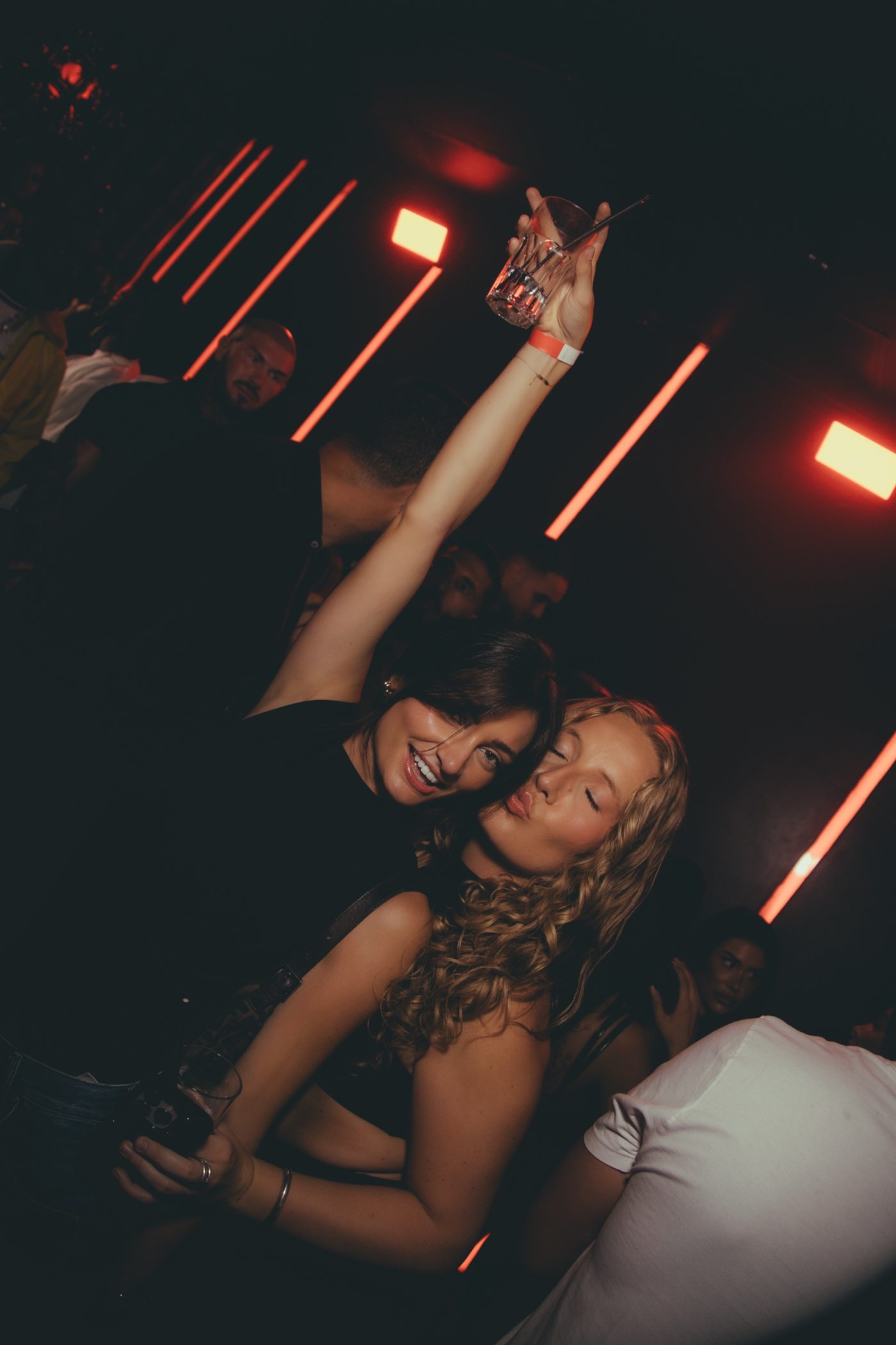 Deux amies qui font la fête, l'une lève son verre, l'autre fait un baiser, tubes rouges au mur