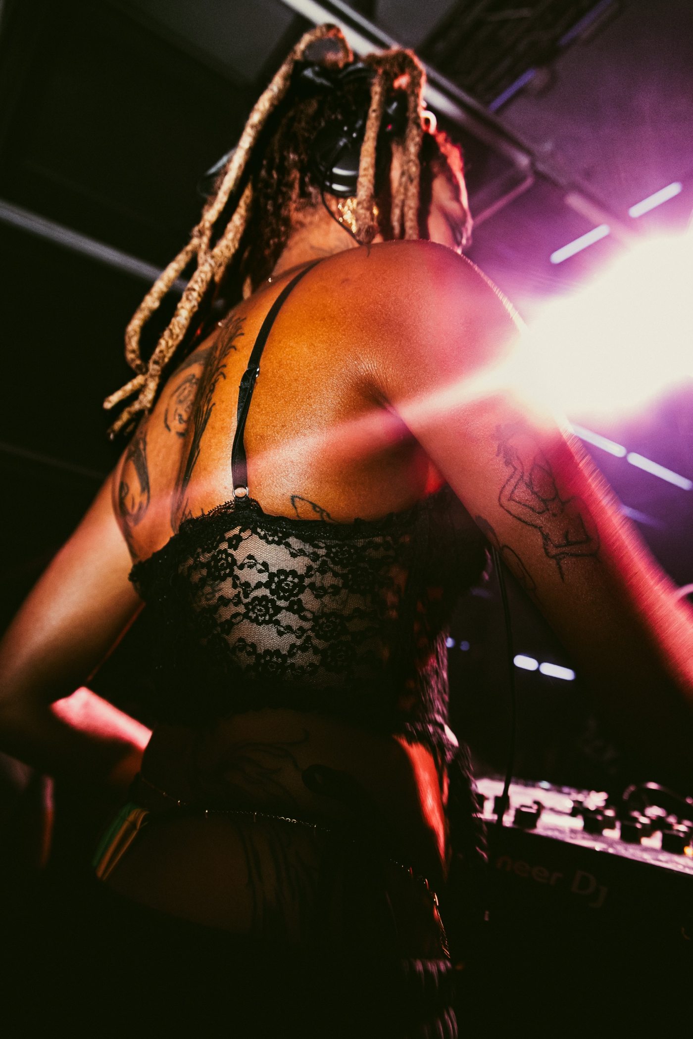 DJ de dos en contre-plongée, brassière en dentelle noire, tatouages et locks, contre-jour violet