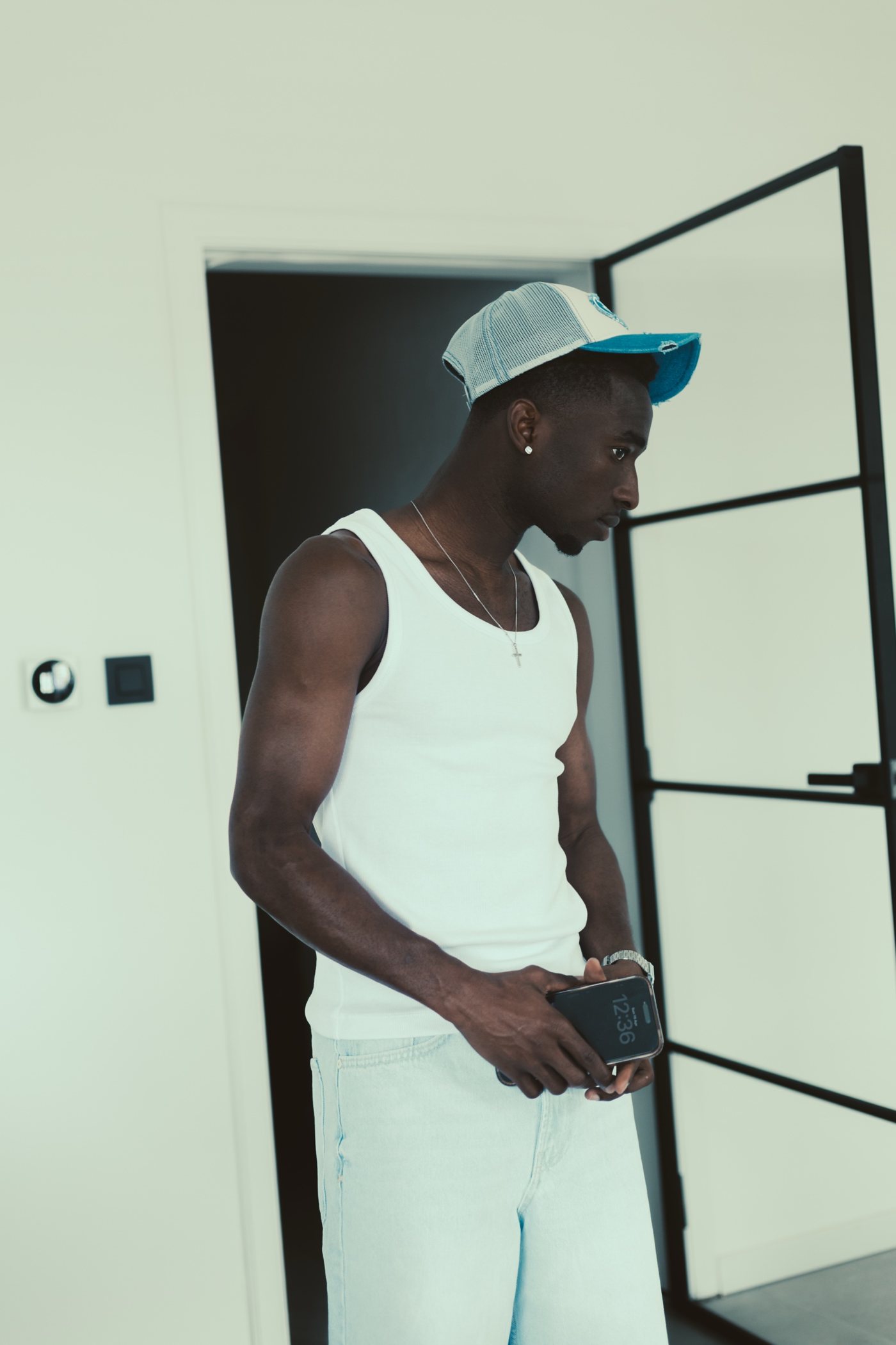 Homme en débardeur blanc et casquette bleue, téléphone à la main, porte vitrée noire