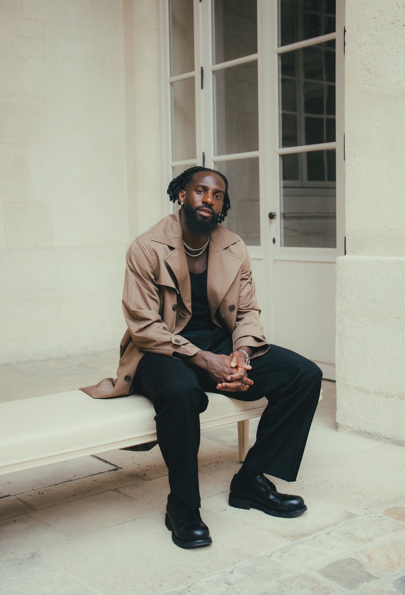 Assis sur un banc dans une cour en pierre, trench beige et pantalon noir