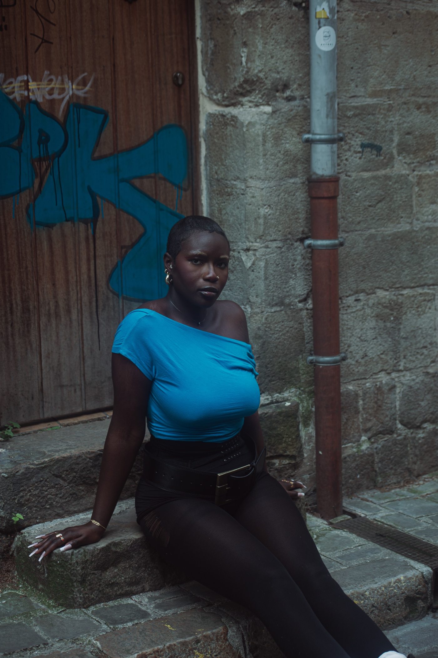 Portrait d'une femme assise sur une marche, haut bleu épaules dénudées, collants noirs, mur de pierre