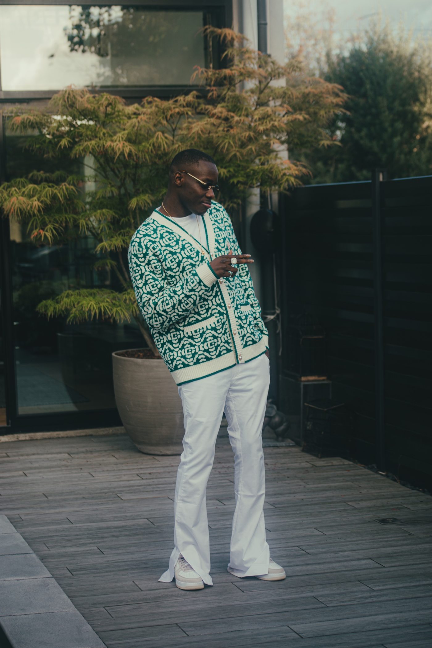 Homme en cardigan vert à motifs et pantalon blanc évasé, lunettes fines, sur une terrasse en bois