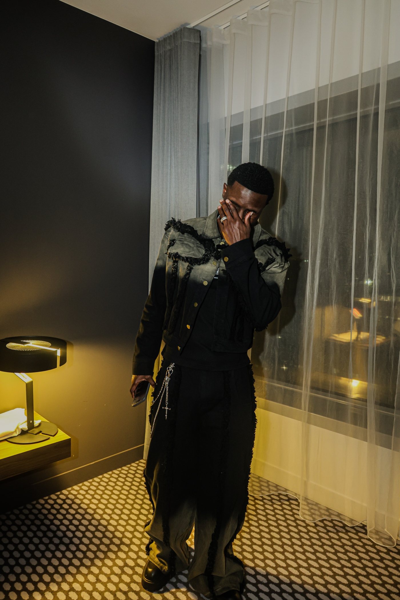 Dans une chambre d'hôtel, veste en jean effilochée, la main sur le visage, lampe jaune